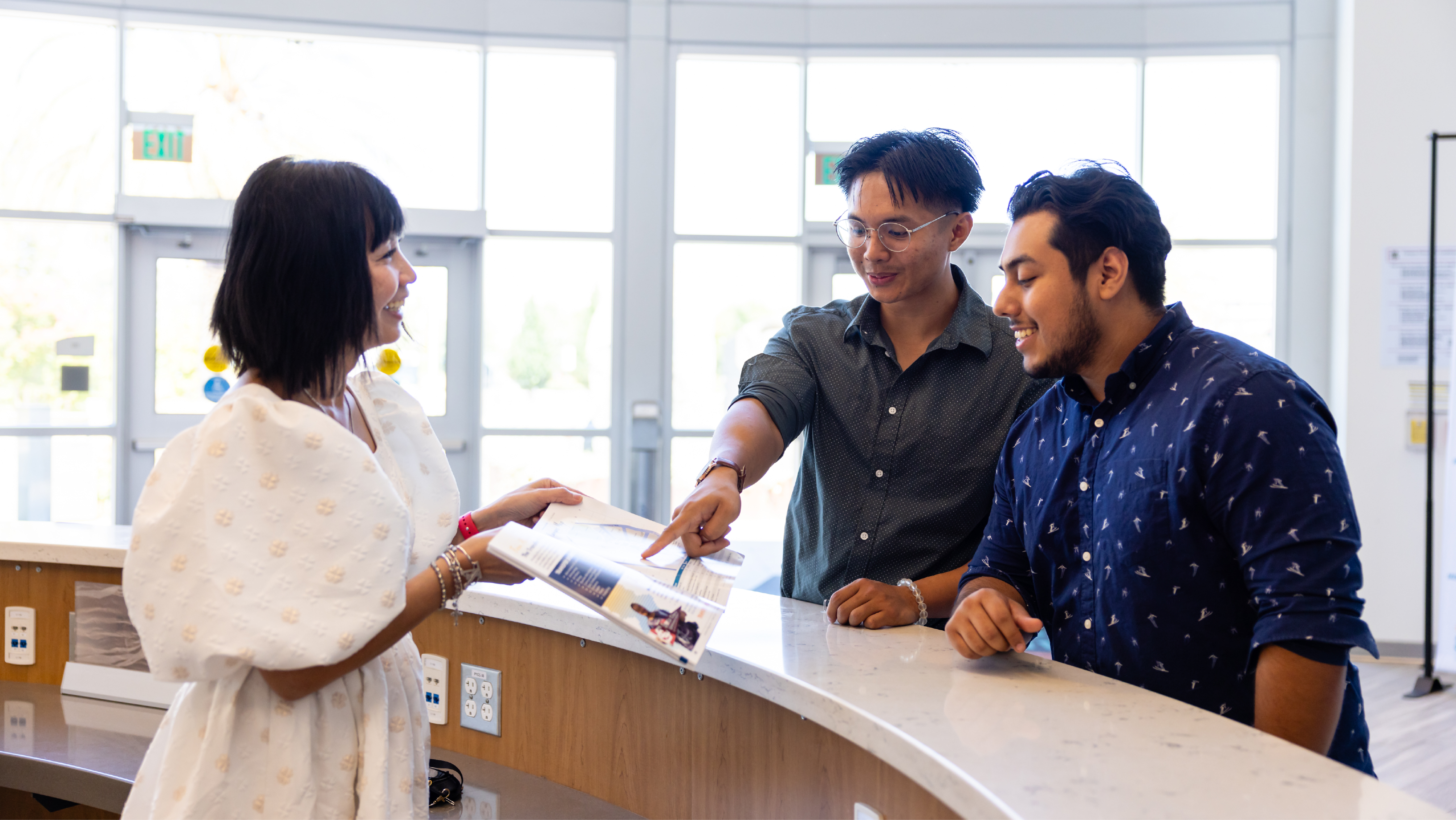Two male students standing at a curved front desk while a staff member in a white dress shows them information inside an open booklet.