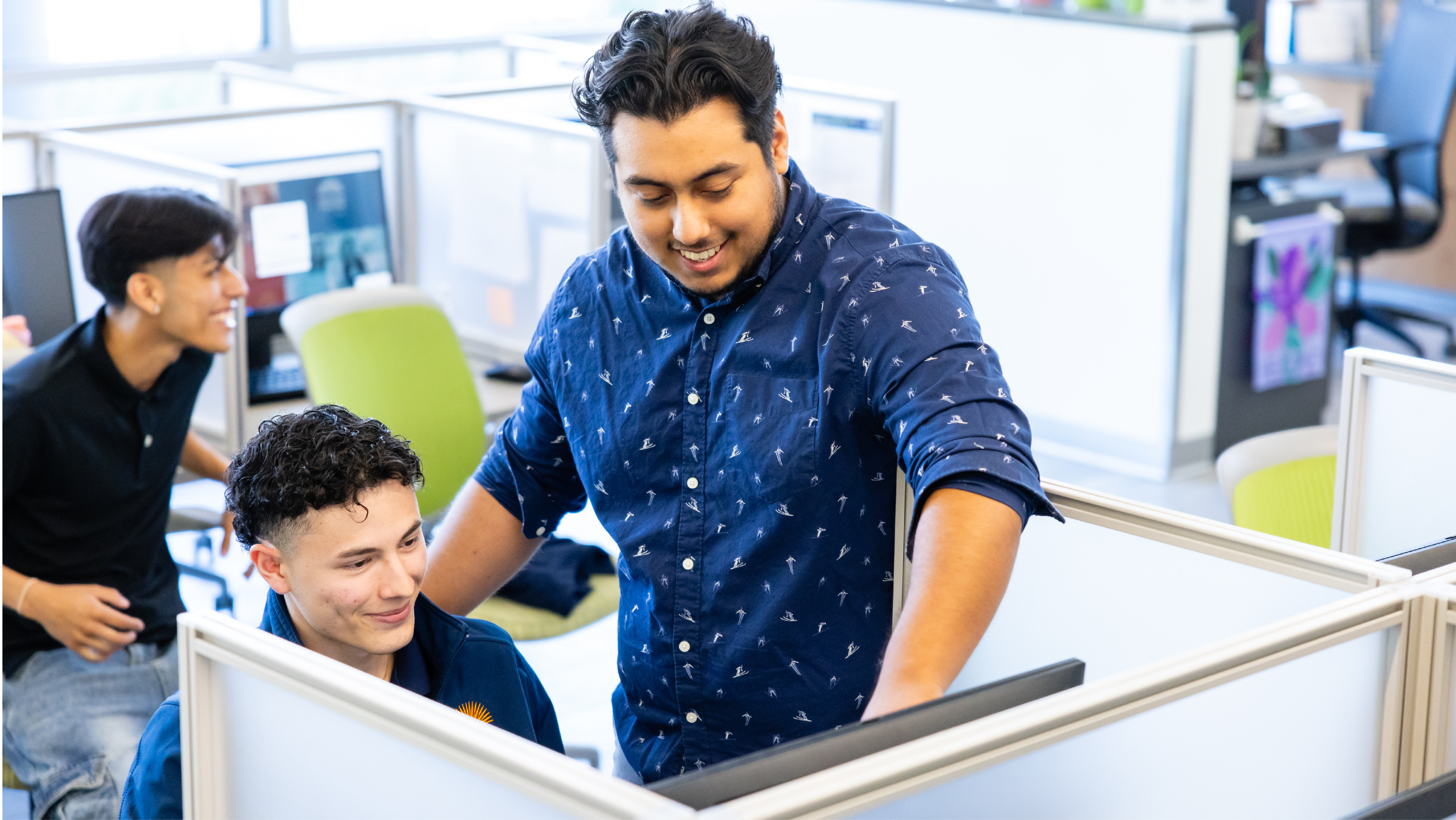 A male staff member in a patterned blue shirt helping a student at a computer station inside a office cubicle setting while another student smiles nearby.