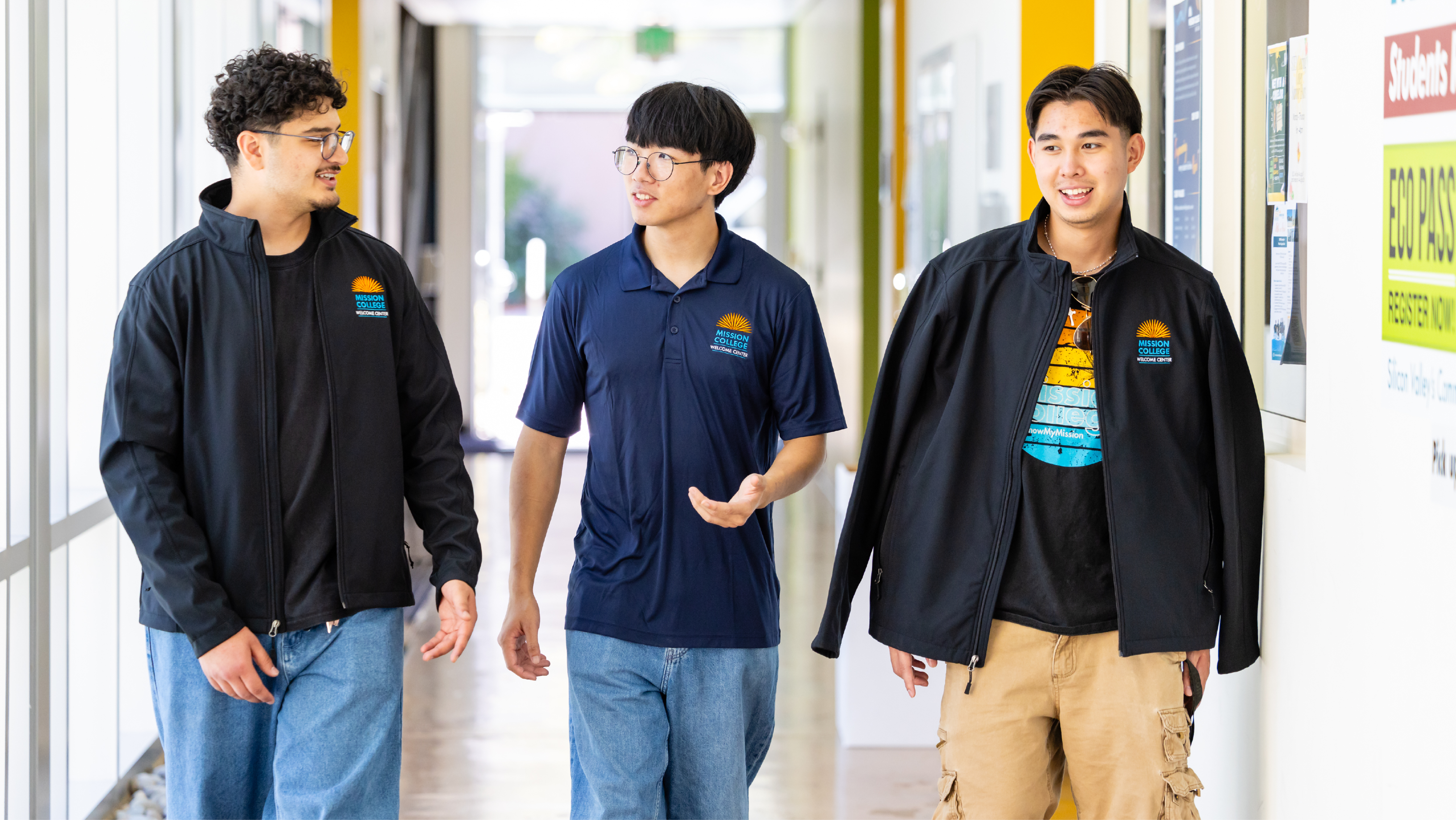 Three male students wearing Mission College Welcome Center apparel walking and talking together down a brightly lit campus hallway.