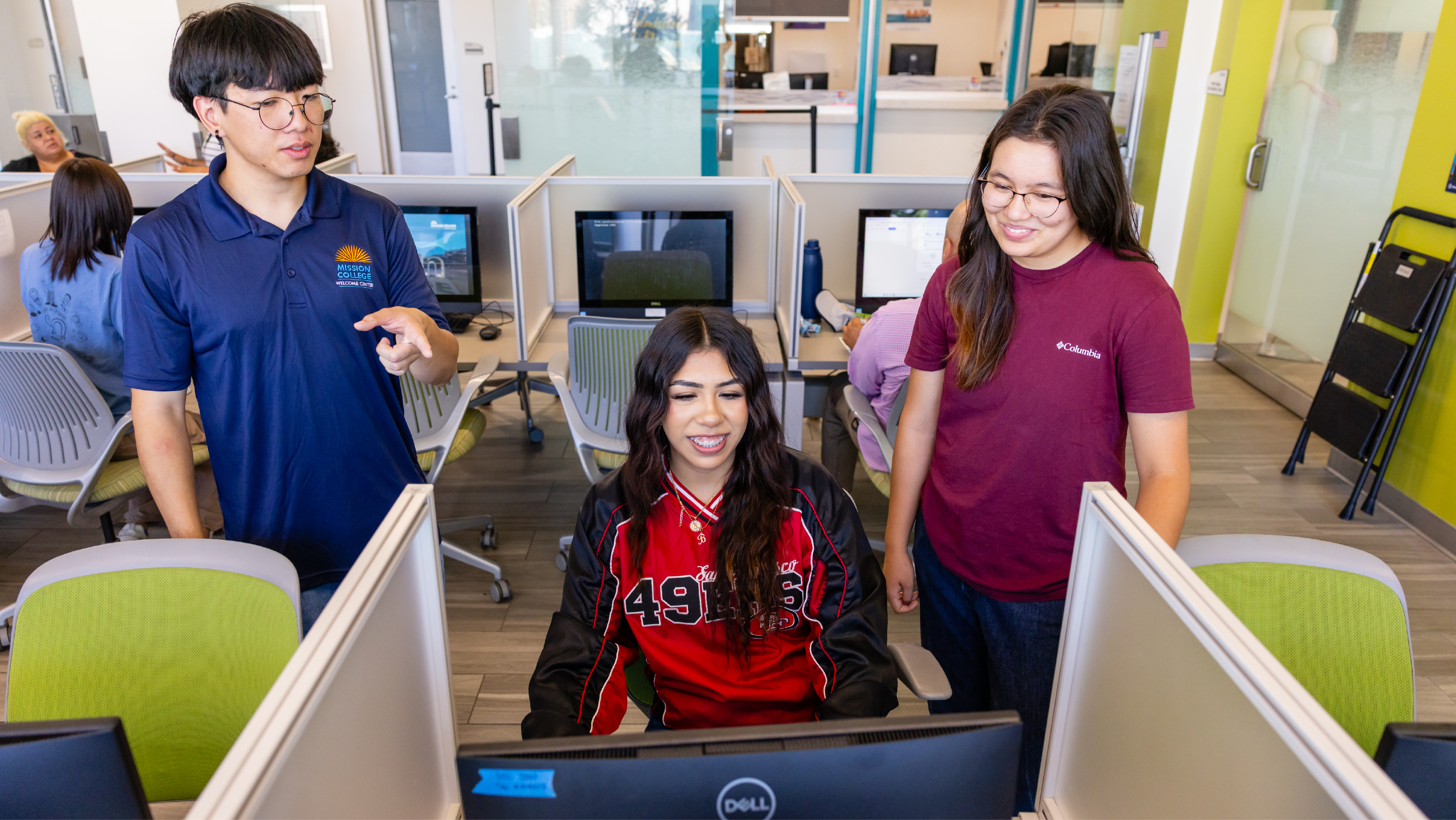 A female student in a red San Francisco 49ers jersey sitting at a computer workstation while a student assistant in a blue polo points to the screen and another student looks on.