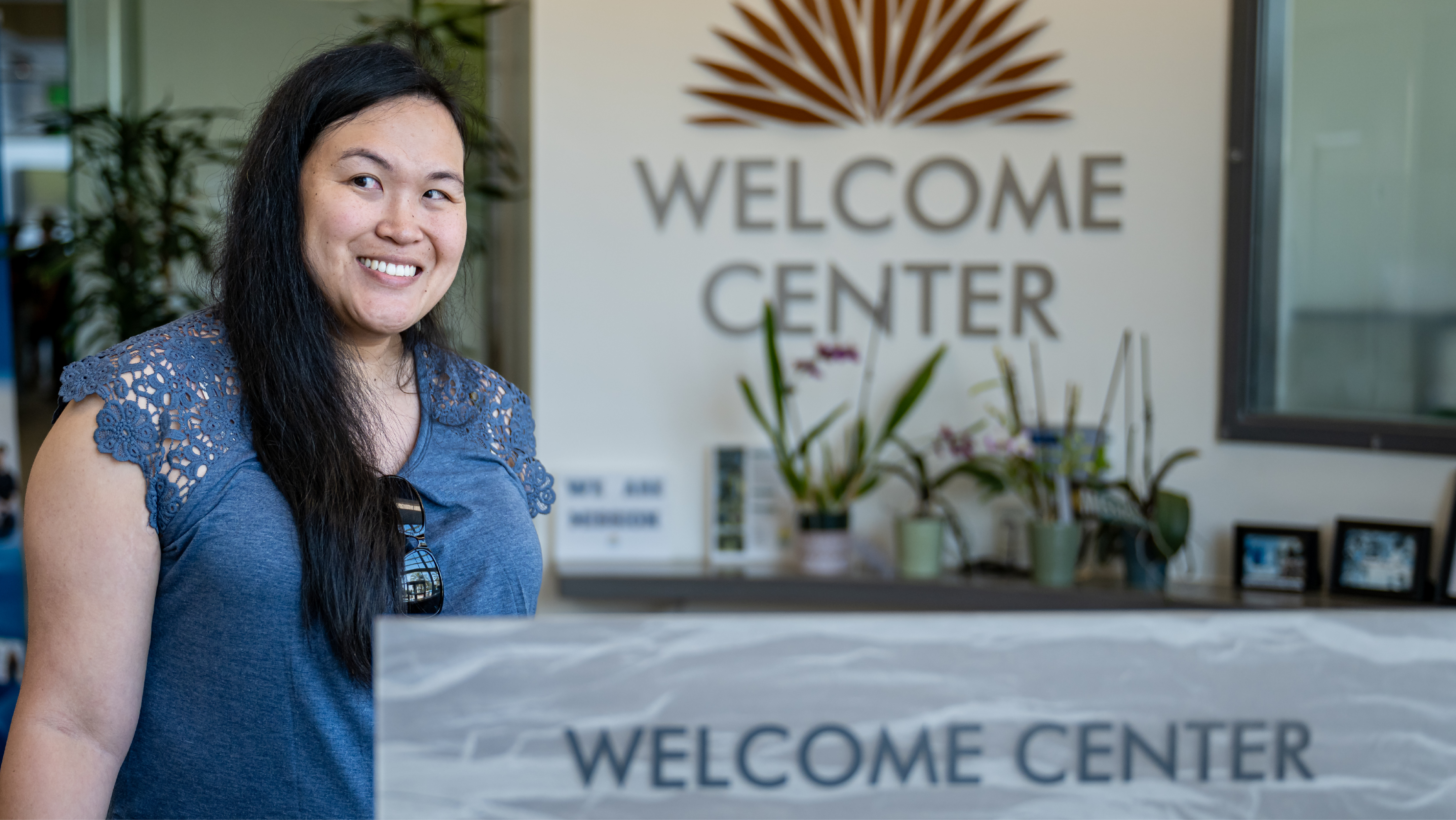 A smiling woman in a blue top standing behind a front desk featuring a prominent "WELCOME CENTER" sign on the wall behind her.
