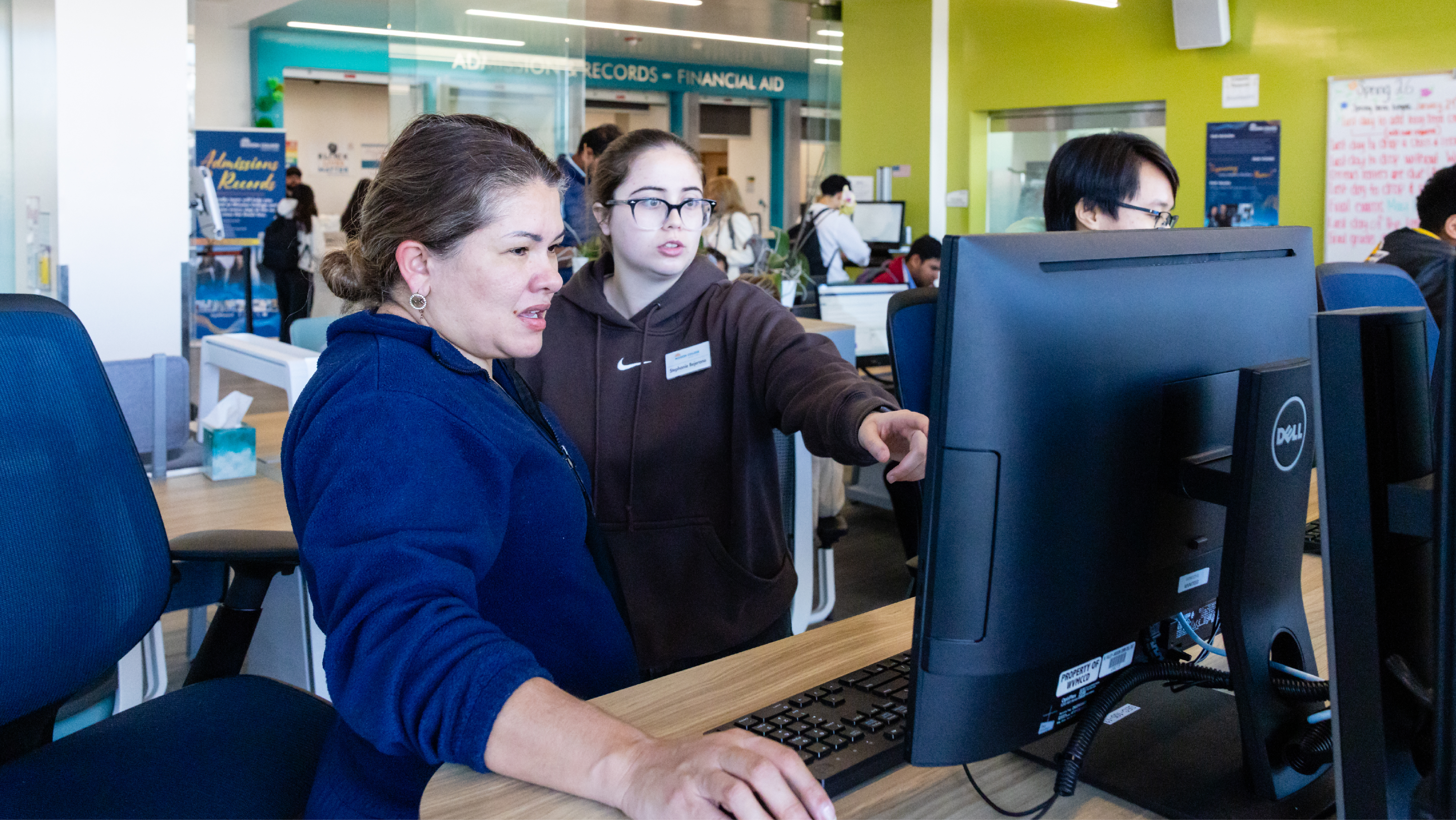 A staff member or student assistant pointing to a computer screen to assist a seated student at a work table in a busy campus center.