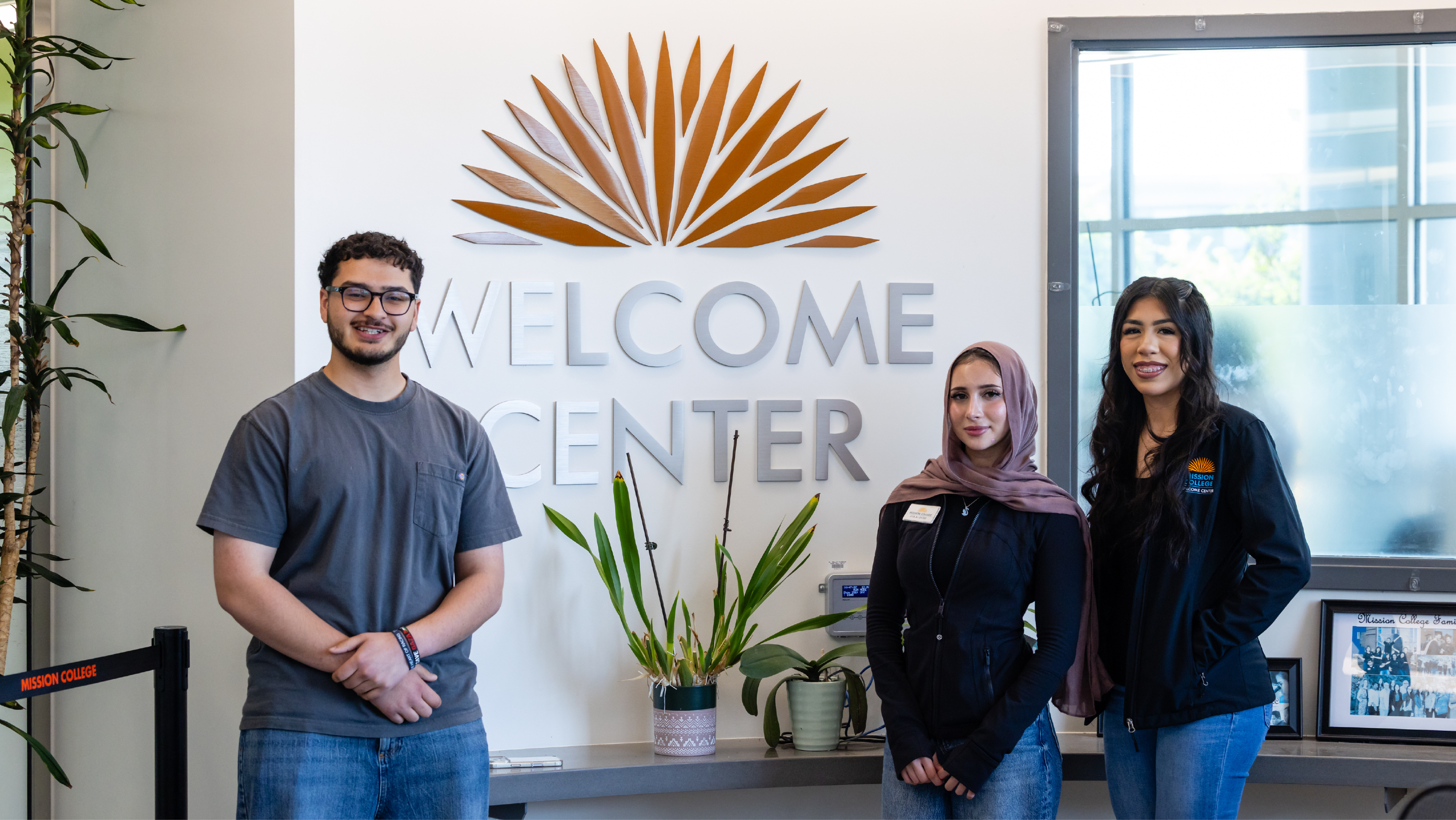 Three student ambassadors posing together indoors in front of a wall featuring the Mission College Welcome Center sign and logo.