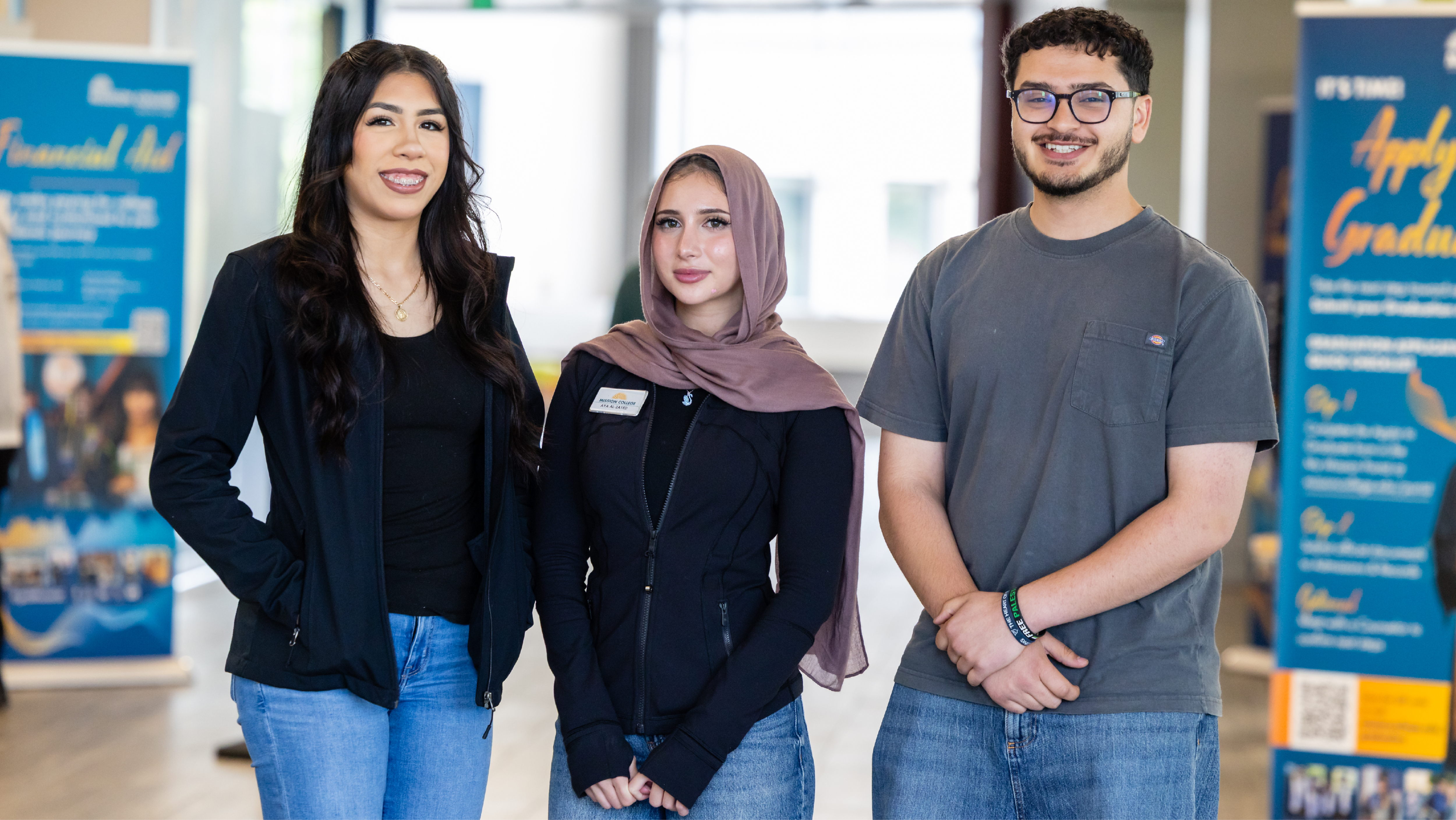Three student ambassadors standing side by side in a lobby area with promotional banners displayed in the background.