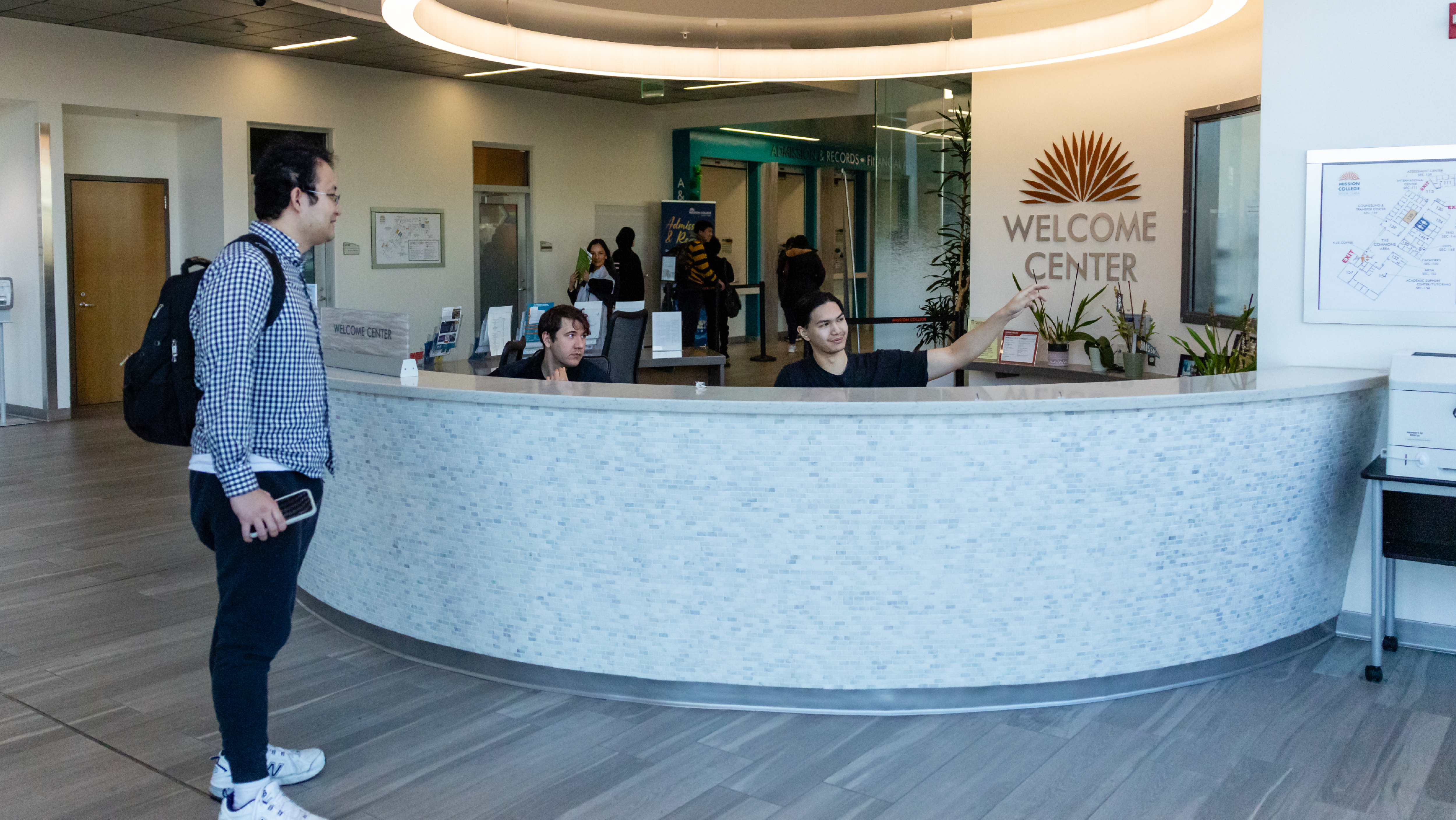 A student with a backpack asking for directions at a large, curved reception desk while a staff member points across the lobby toward the Welcome Center.