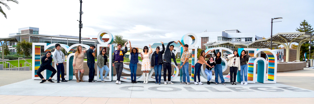 Group of students in jeans and sweatshirts pose in front of the Welcome Center on campus.