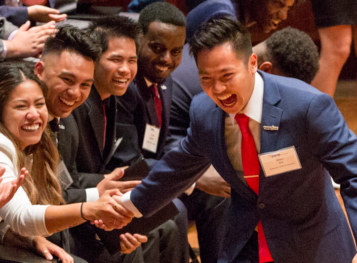 Asian-American male student wears a blue suit and tie. He runs by a row of seated students giving them "high-fives." at their commencement ceremony for Year Up.