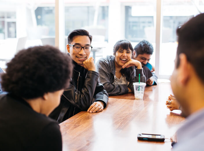 Group of young adults in business casual attire sit around a table in a modern office conference room.