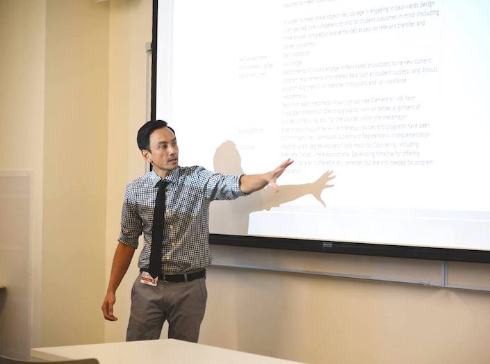 Dr. Danny Nguyen, a man in a long-sleeved checked button down shirt, gestures toward a marker board with writing on it. He is of Vietnamese decent, slight, and has sharp attractive features and short black hair.