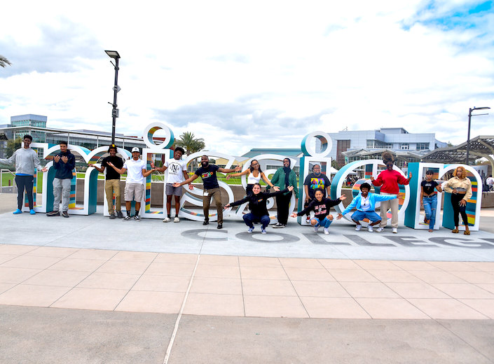 Group of Umoja students pose in front of Umoja sign (big block letters) on campus in light teal, yellow, orange, and white.