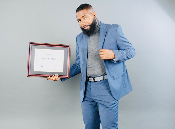 Roshawn Walter, a youngish man of African-American descent wears a fitted blue suit and holds an award. He has a full beard.