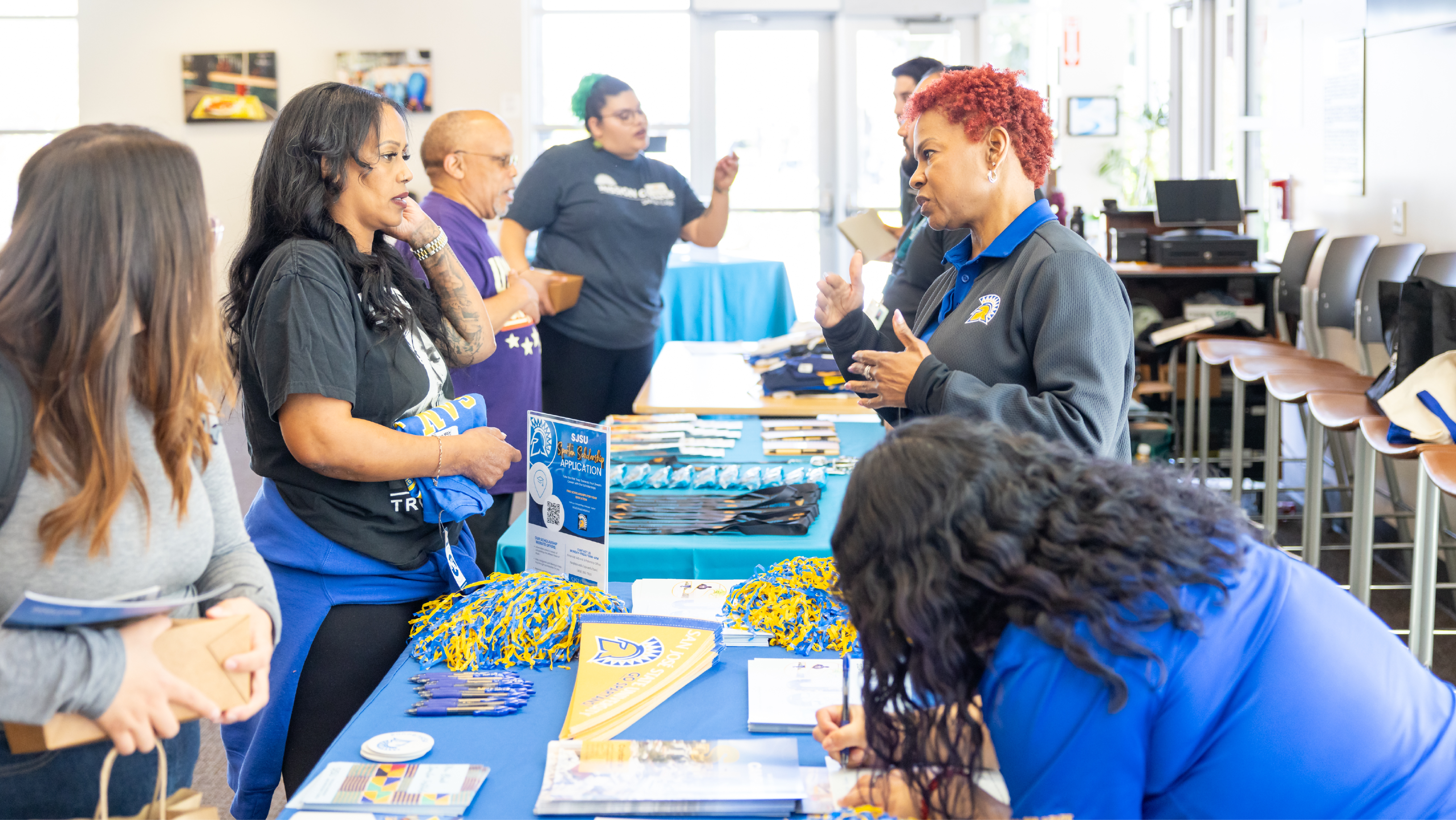 Representative speaking with students and guests gathered around a San José State University information table loaded with pennants, brochures, and flyers.