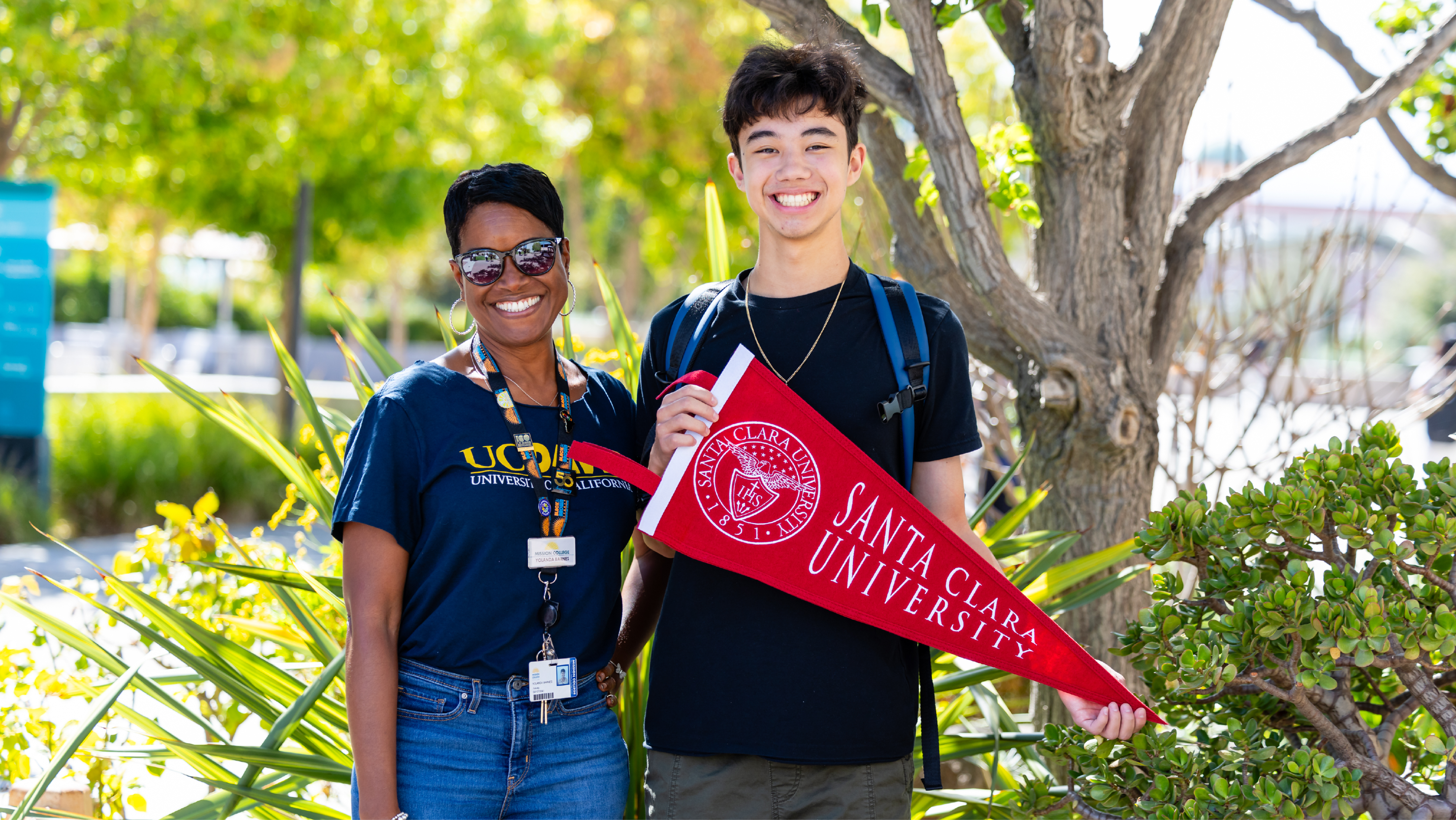 A Mission College staff member and a student posing together outdoors while holding a red Santa Clara University pennant.