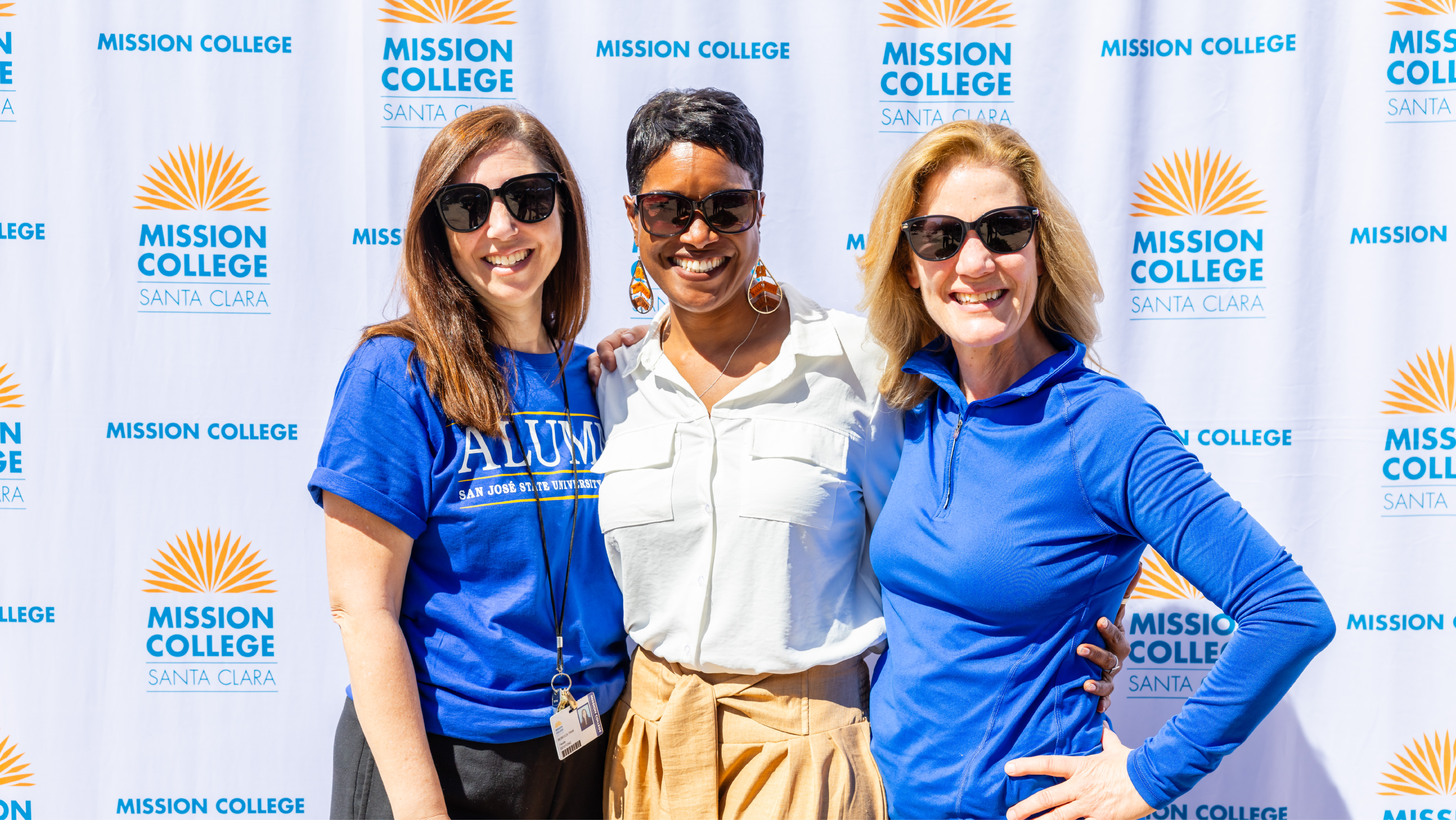 Three Mission College staff members smiling together in front of a branded photo backdrop.