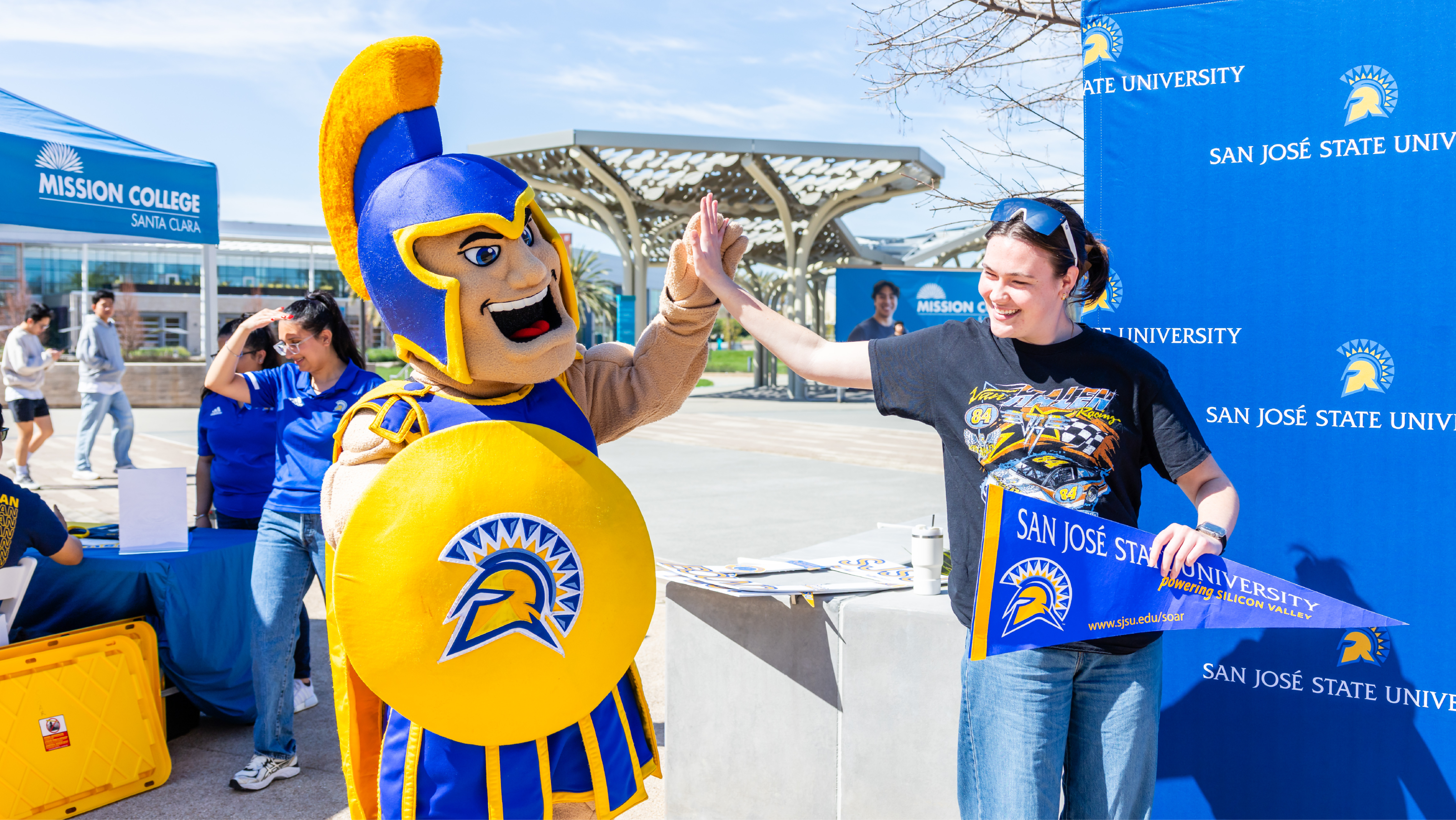 A student giving a high five to the San José State University Spartan mascot while holding a university pennant outdoors.
