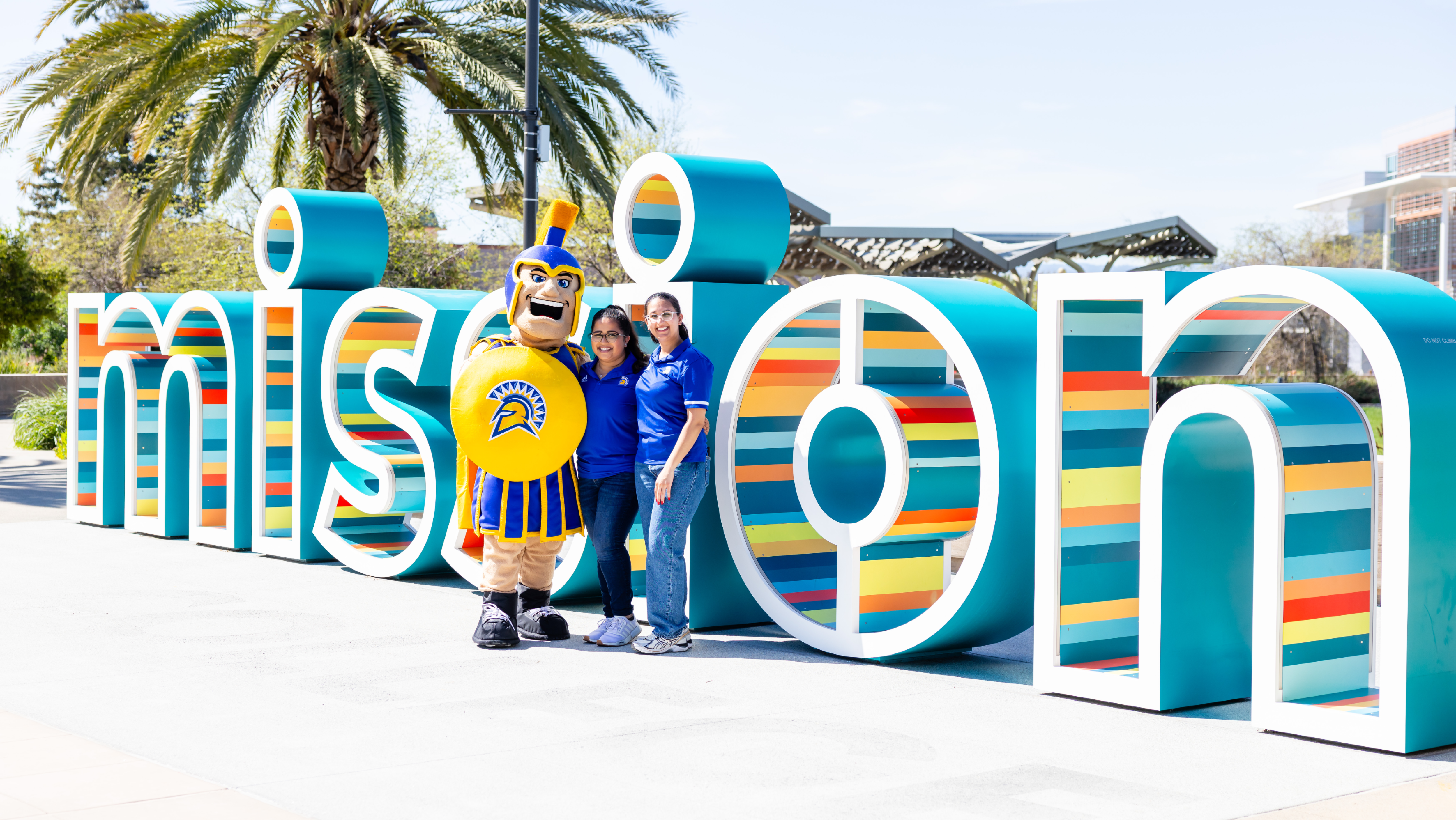 Two staff members and the SJSU Spartan mascot posing together in front of the large outdoor Mission College campus sign.