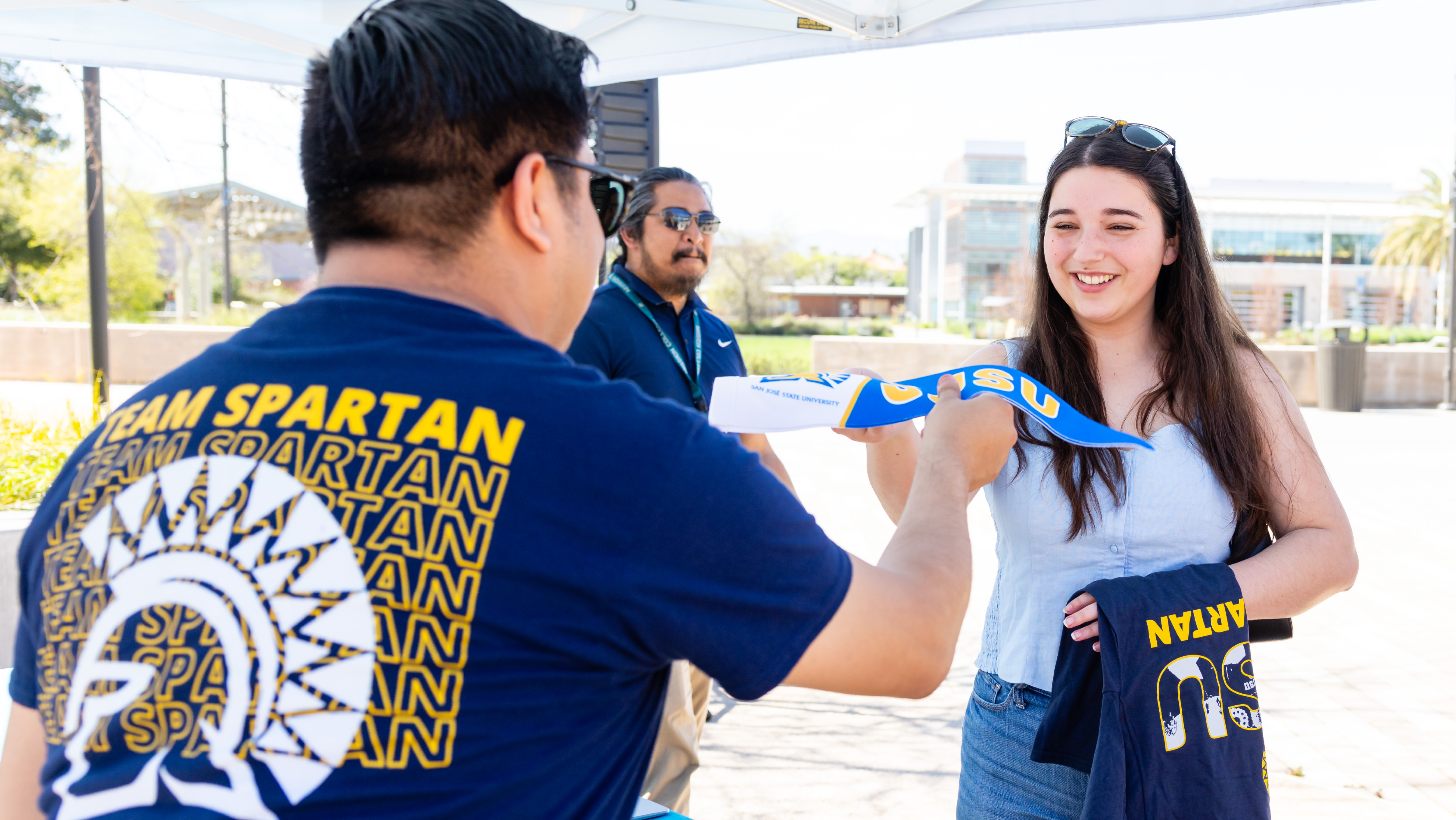 A staff member at an event table handing a blue and yellow San José State pennant to a smiling student.
