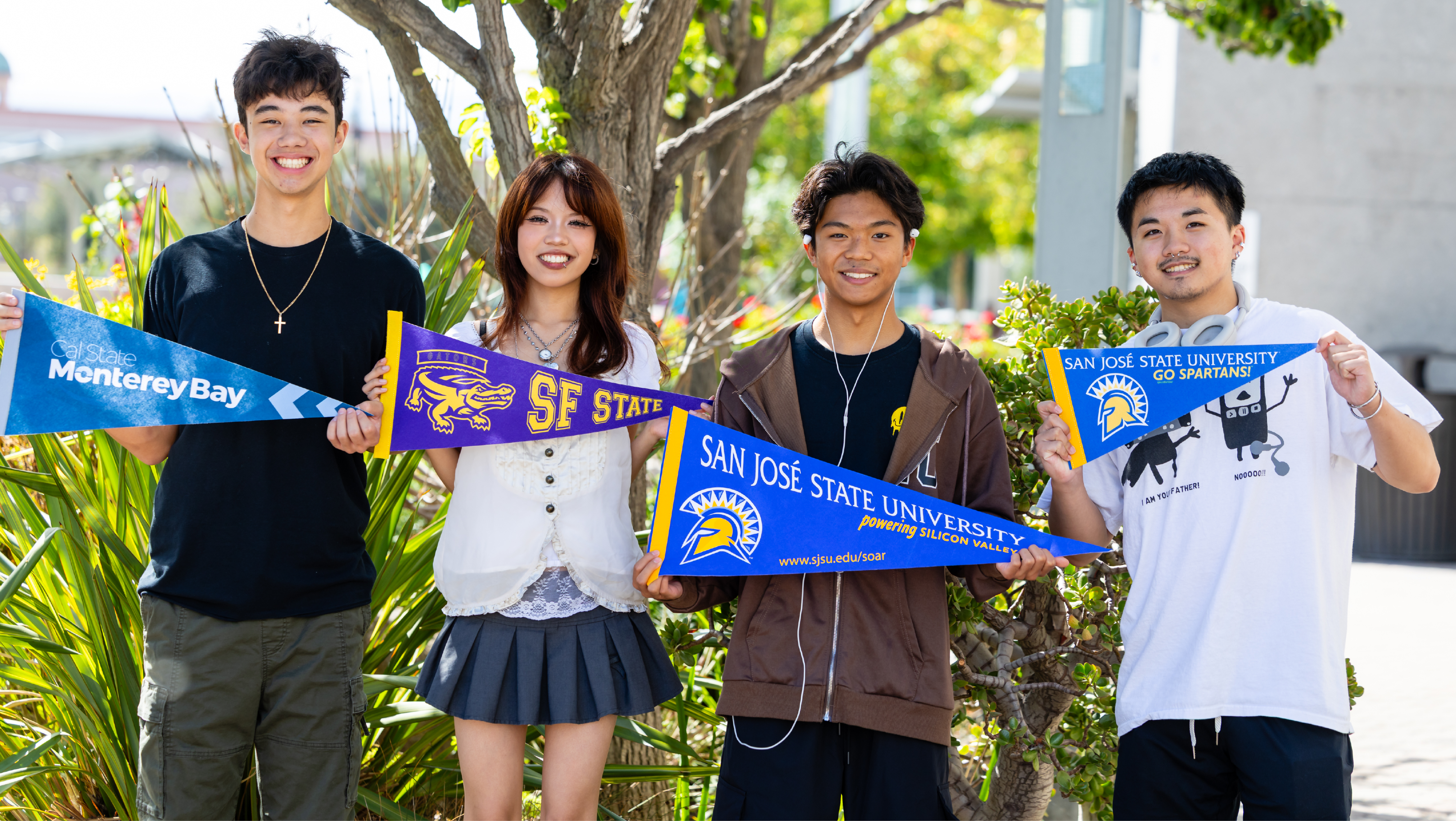 Four students standing together outdoors holding university pennants for Cal State Monterey Bay, SF State, and San José State.