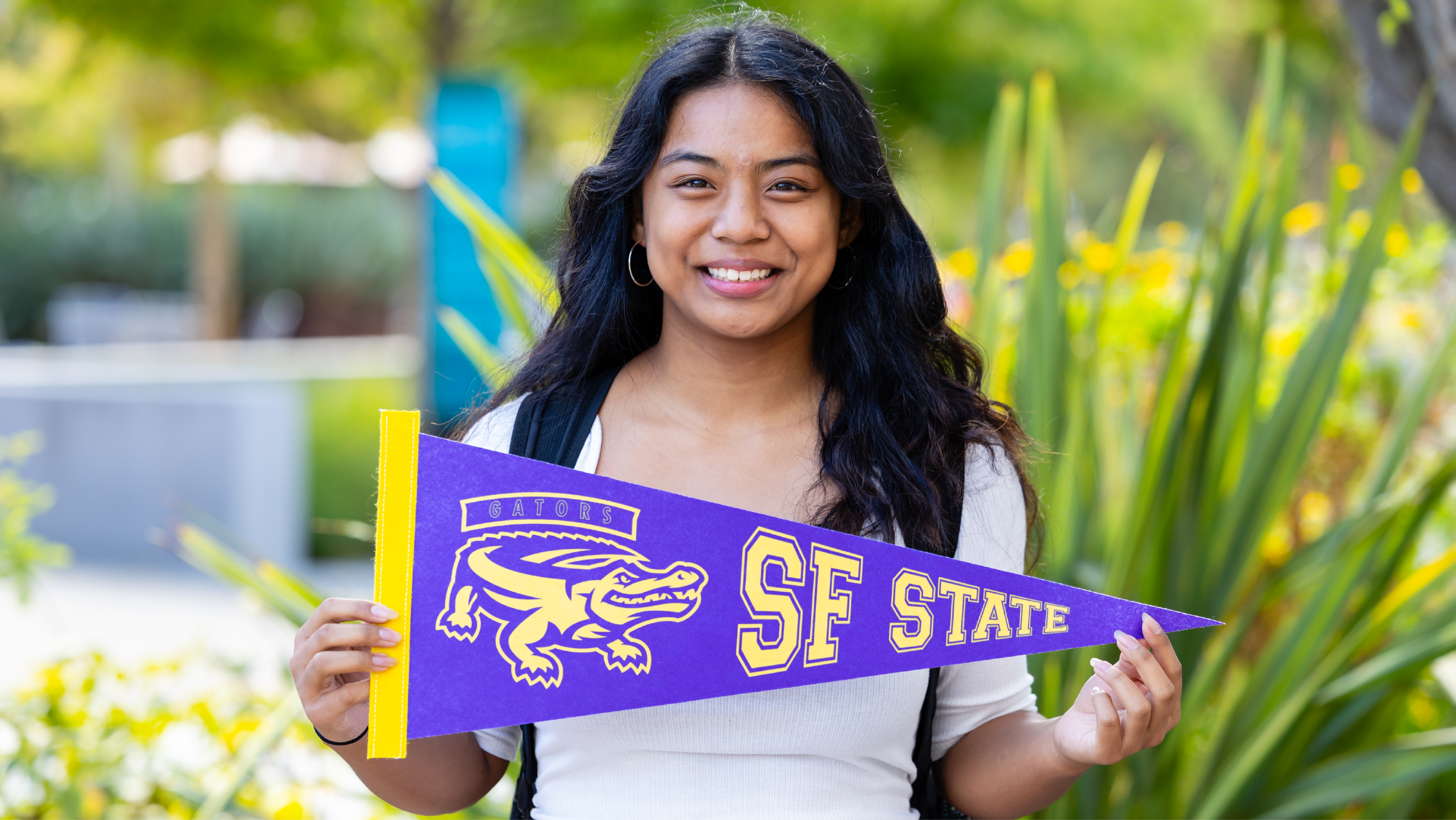 A smiling student holding up a purple SF State Gators pennant outdoors.