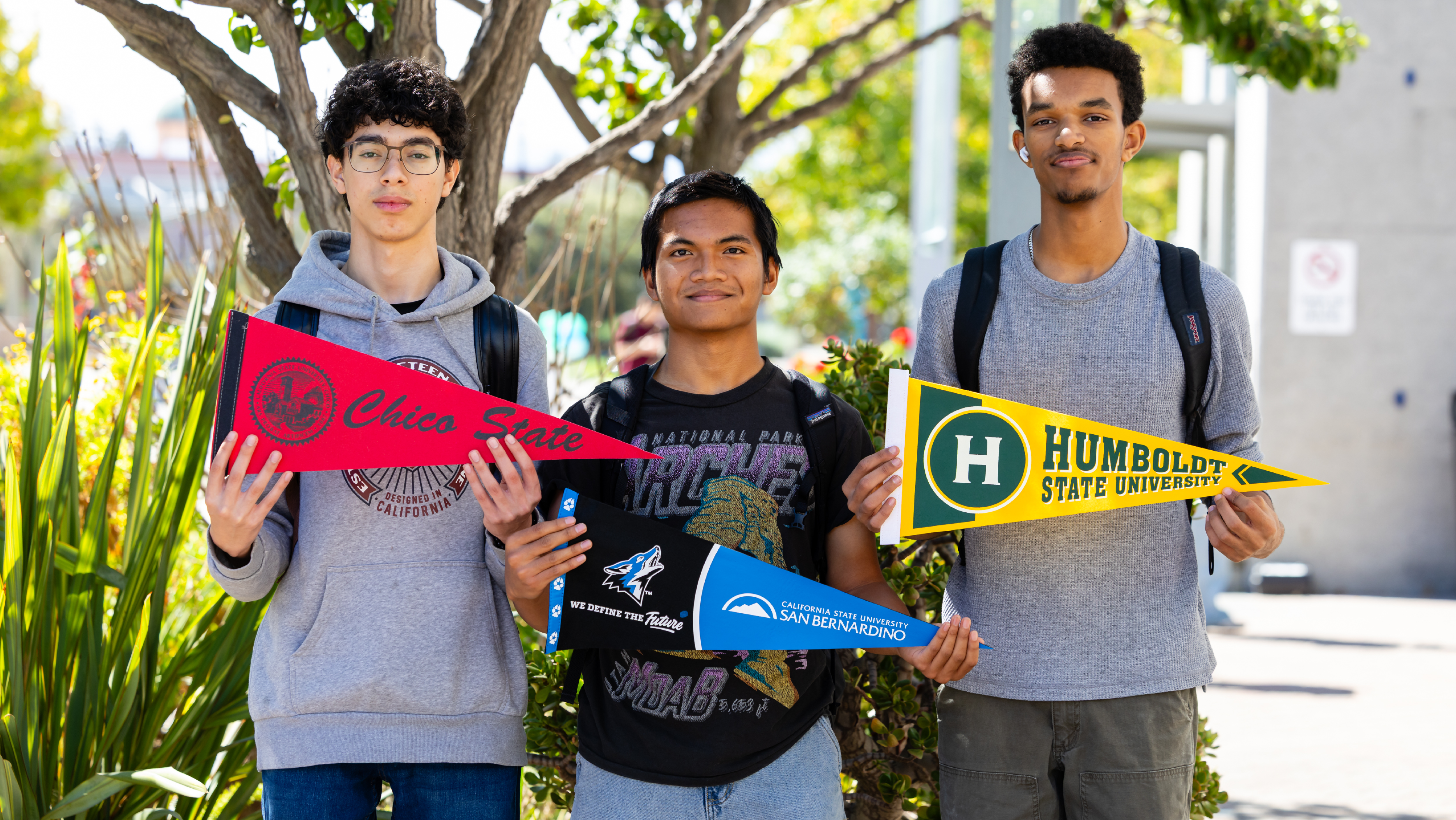 Three students standing side-by-side outdoors holding pennants for Chico State, CSU San Bernardino, and Humboldt State University.