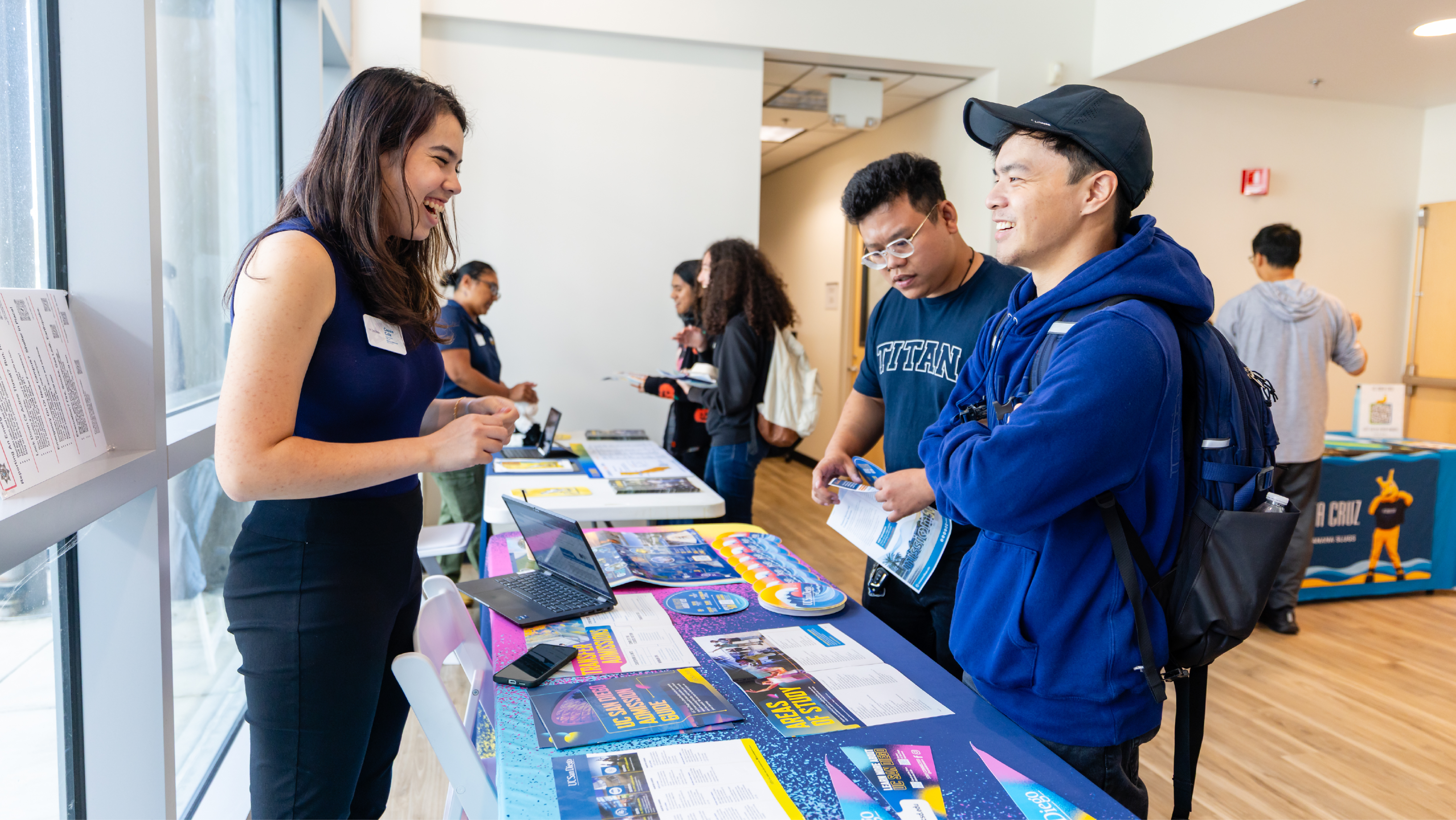 A university representative speaking with two male students at an indoor transfer information booth.