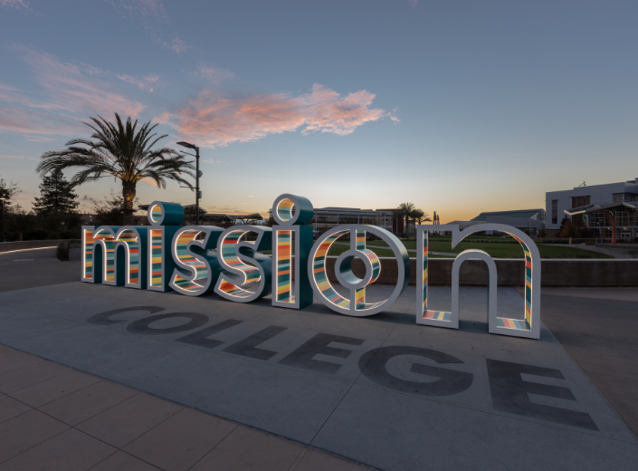 The Mission College entrance sign stands at the front of campus in Santa Clara, California, with palm trees, campus buildings, and a sunset sky in the background. The image represents Mission College, one of five community colleges selected nationwide to partner with Intel on a new artificial intelligence education pilot for transitioning foster youth.