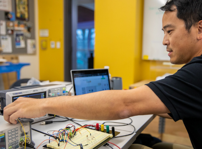 A Mission College Mechatronics student works hands-on at an engineering lab bench, fine-tuning testing equipment wired to a breadboard circuit prototype.