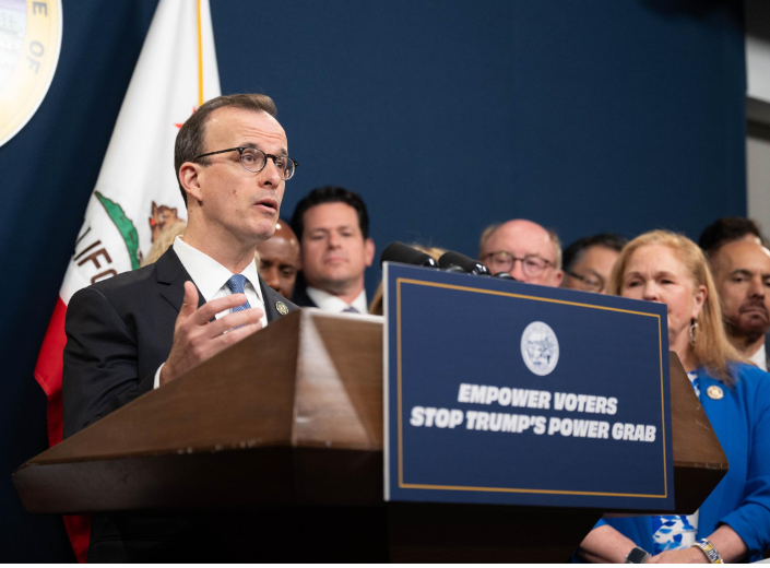 California Assemblymember Marc Berman speaks at a wooden podium featuring a sign that reads "Empower Voters Stop Trump’s Power Grab," flanked by state leaders, colleagues, and the California state flag in the background.
