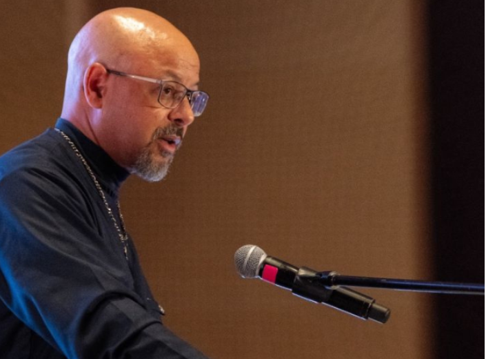 Dr. Chike Akua, wearing glasses and dark attire, speaks into a microphone at a podium during a presentation.