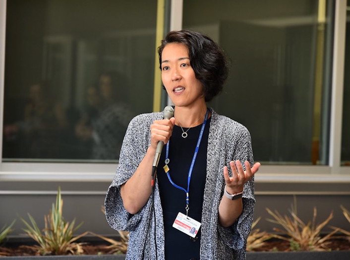 Chigusa, a Japanese-American woman with wavy chin-length hair holds a microphone and speaks at an event.