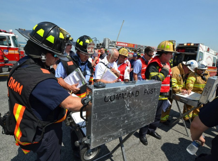First responders, including an incident commander and planning section specialist wearing high-visibility vests and helmets, observe operations near fire trucks.