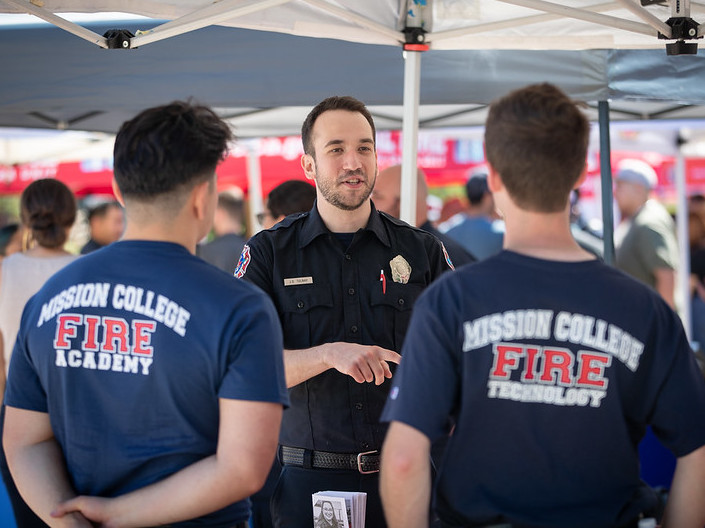 A professional firefighter speaks to two Firefighter cadets in dark blue t-shirts with "Mission College Fire Department" on the back. 