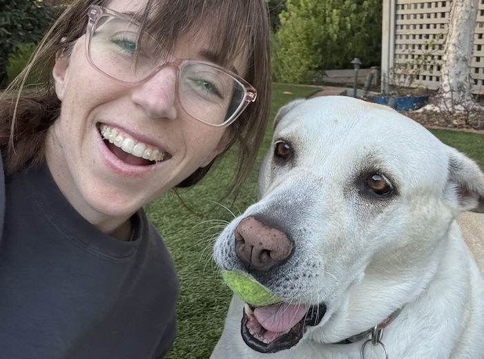 Brittany Gunnel, a white woman with long brown hair poses with her white lab. The dog holds a tennis ball in it's mouth.
