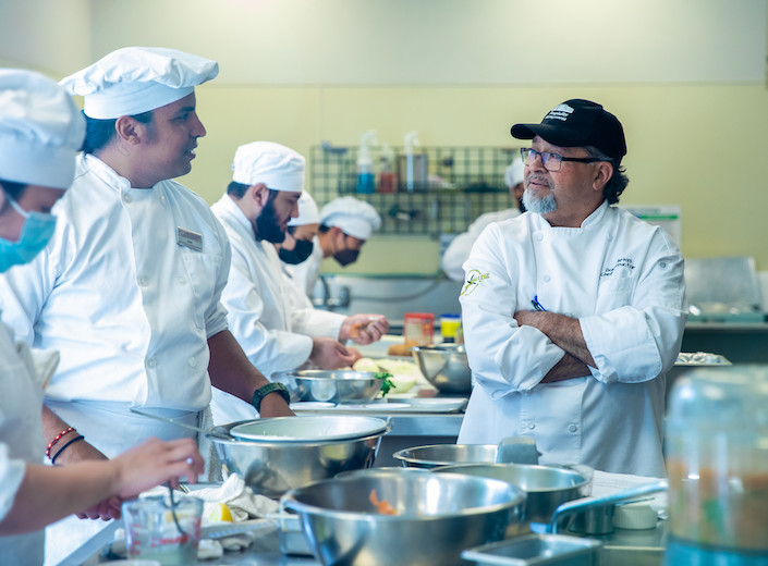 Professor Arias Dennis instructs two students in chef white uniforms with hats on a recipe.