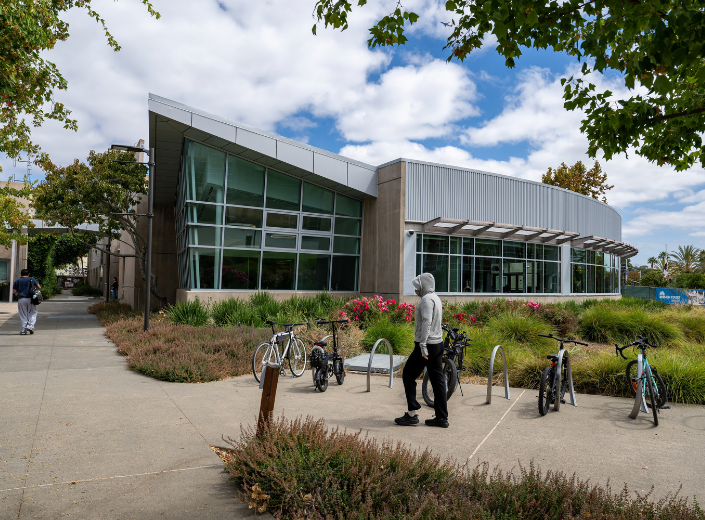 Exterior view of the Mission College Library & Learning Commons showing a curved, modern glass facade, bike racks, and landscaped concrete walkways under a partly cloudy sky.