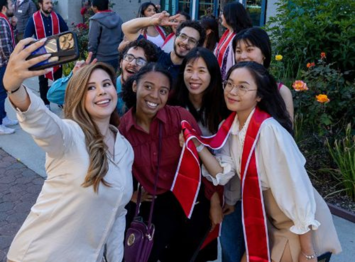 Smiling students pose for selfie at commencement. They are young women of various ethnic backgrounds and one young man with dark hair and glasses in the rear.