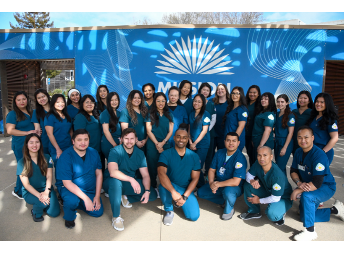 A group of Mission College nursing students wearing teal scrubs pose together outdoors in front of a blue Mission College mural. The diverse cohort smiles for the camera, representing the college's Associate Degree in Nursing program.