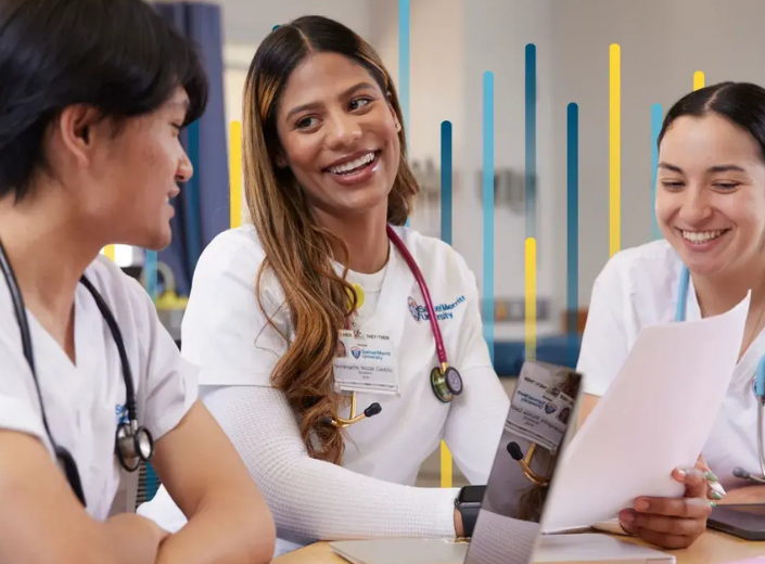 Three nursing students in white clinical uniforms collaborate around a table, smiling as they review documents together during a classroom discussion.