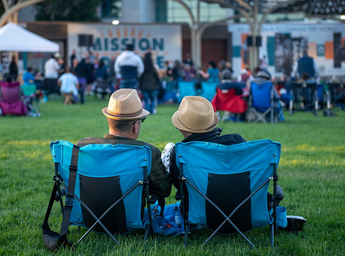 A couple in matching tan hats is seated (pictured from the back) in the Central Plaza and listening to a concert.