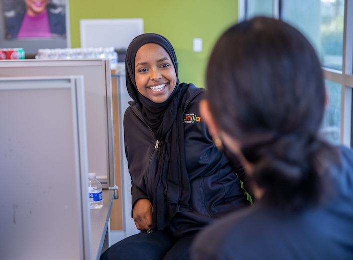 Nasra Mohamed in the Welcome Center. She is a smiling young woman with a dark headscarf and golden-brown skin. She wears a black Umoja jacket.