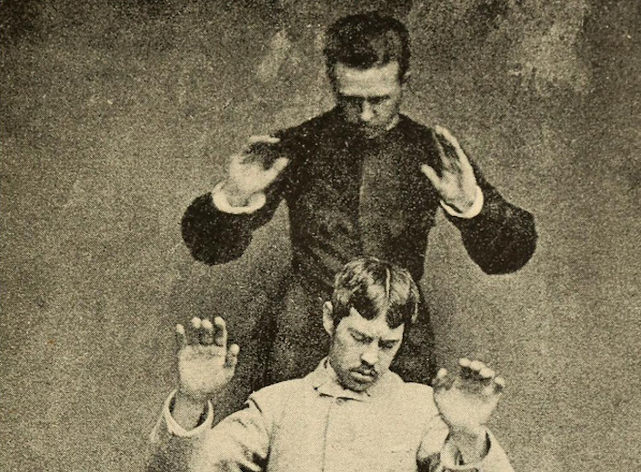 An old photo of a man with his hands up in the air. He is standing behind another man who looks down with his eyes closed in concentration.